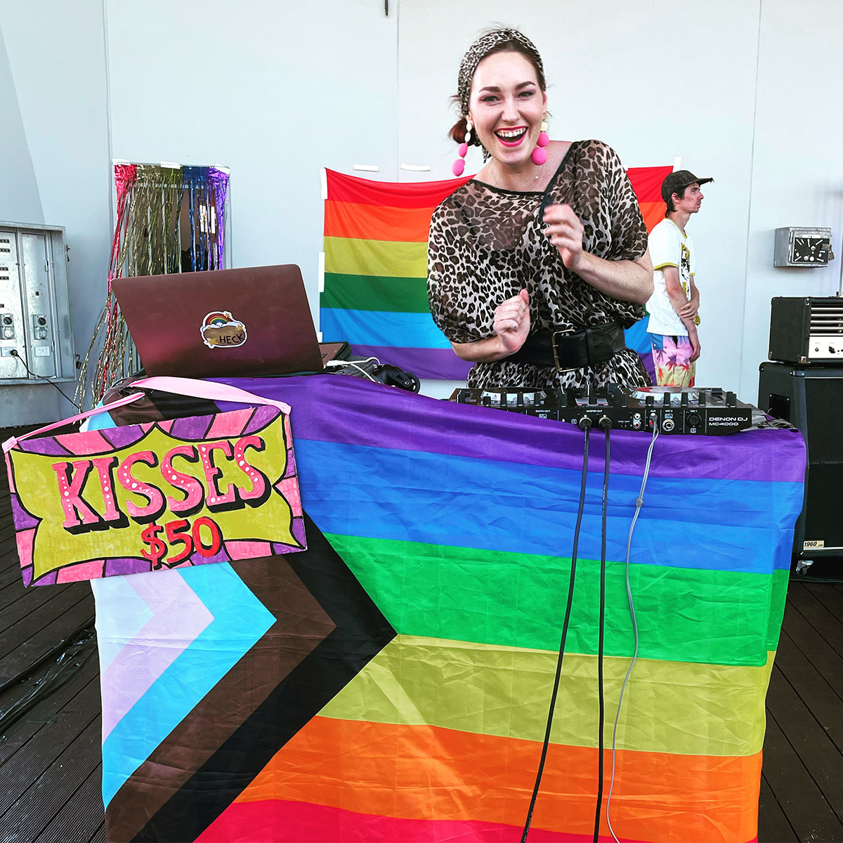 A fun pic of Ginger LaMinge wearing a leopard print in front of the Progress Pride Flag and a sign that says 'KISSES $50'