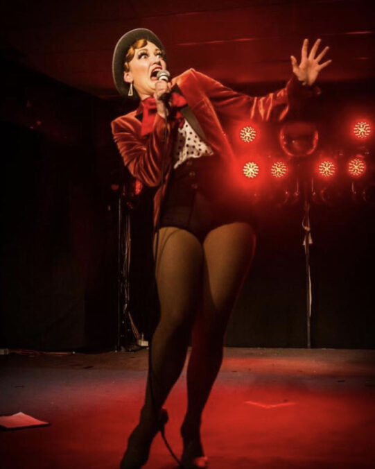 Ginger LaMinge wearing a red velvet jacket and performing on stage with red lighting and an outrageous expression.