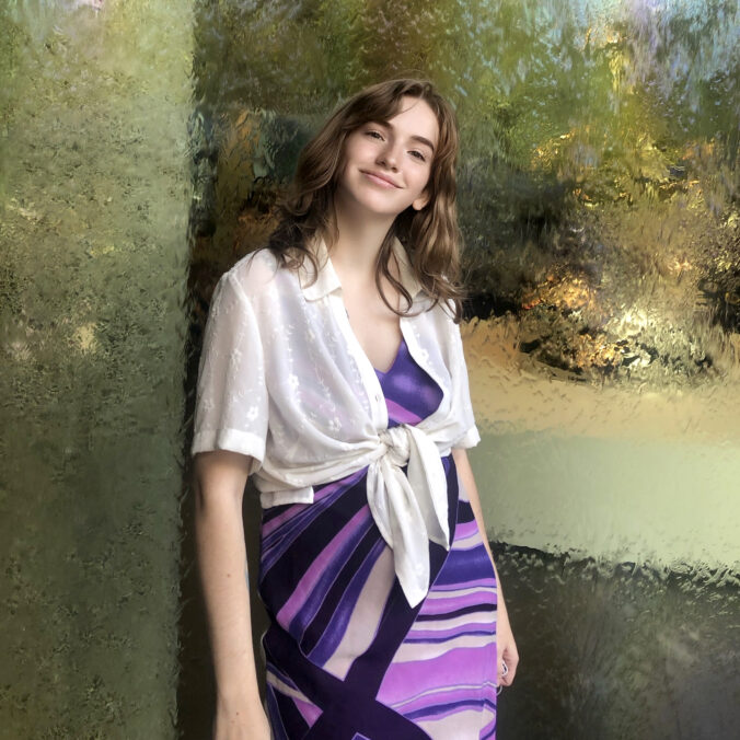 Picture of Caroline smiling shyly, wearing a purple dress, white shawl and standing against a gold shimmering background.