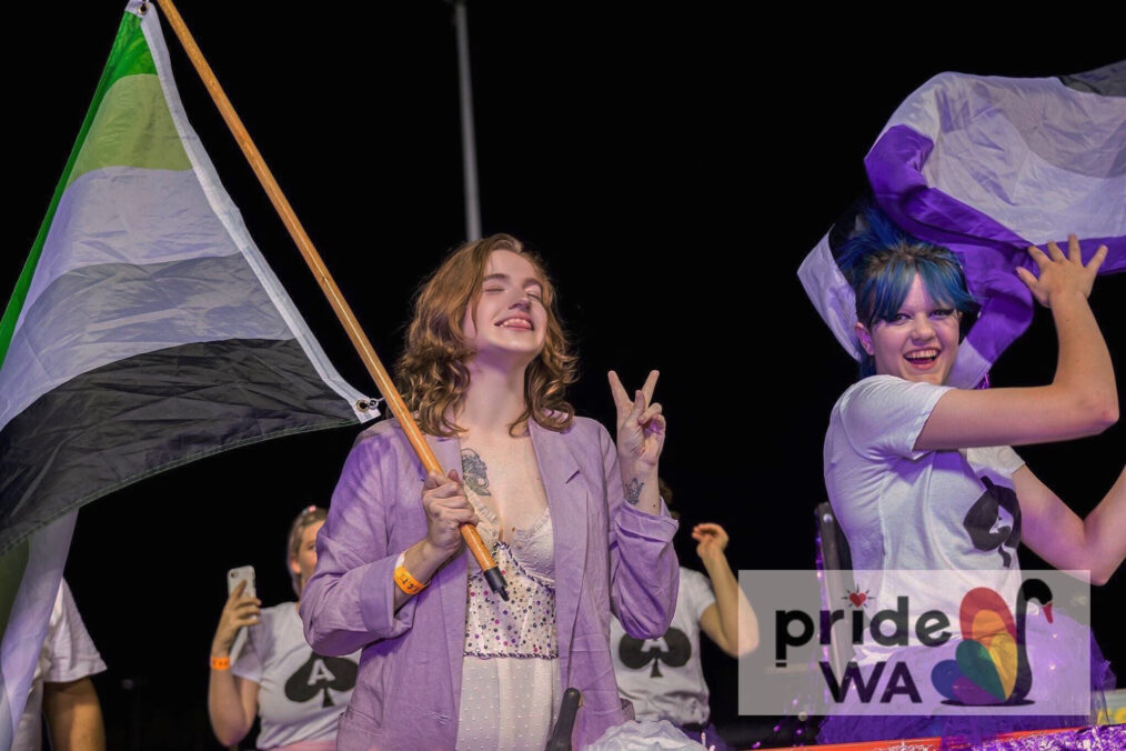 Photo showing Caroline marching in the WA Pride Parade grinning, making the peace sign with her fingers and holding the aromantic flag.